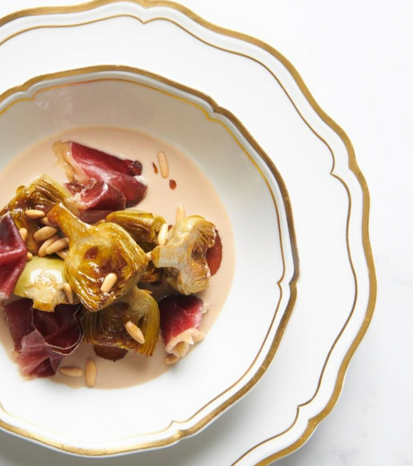 A gourmet dish of caramelized artichokes, cured meat, pine nuts, and creamy sauce is artfully presented on a white Polka Gold Dinnerware plate by Raynaud.
