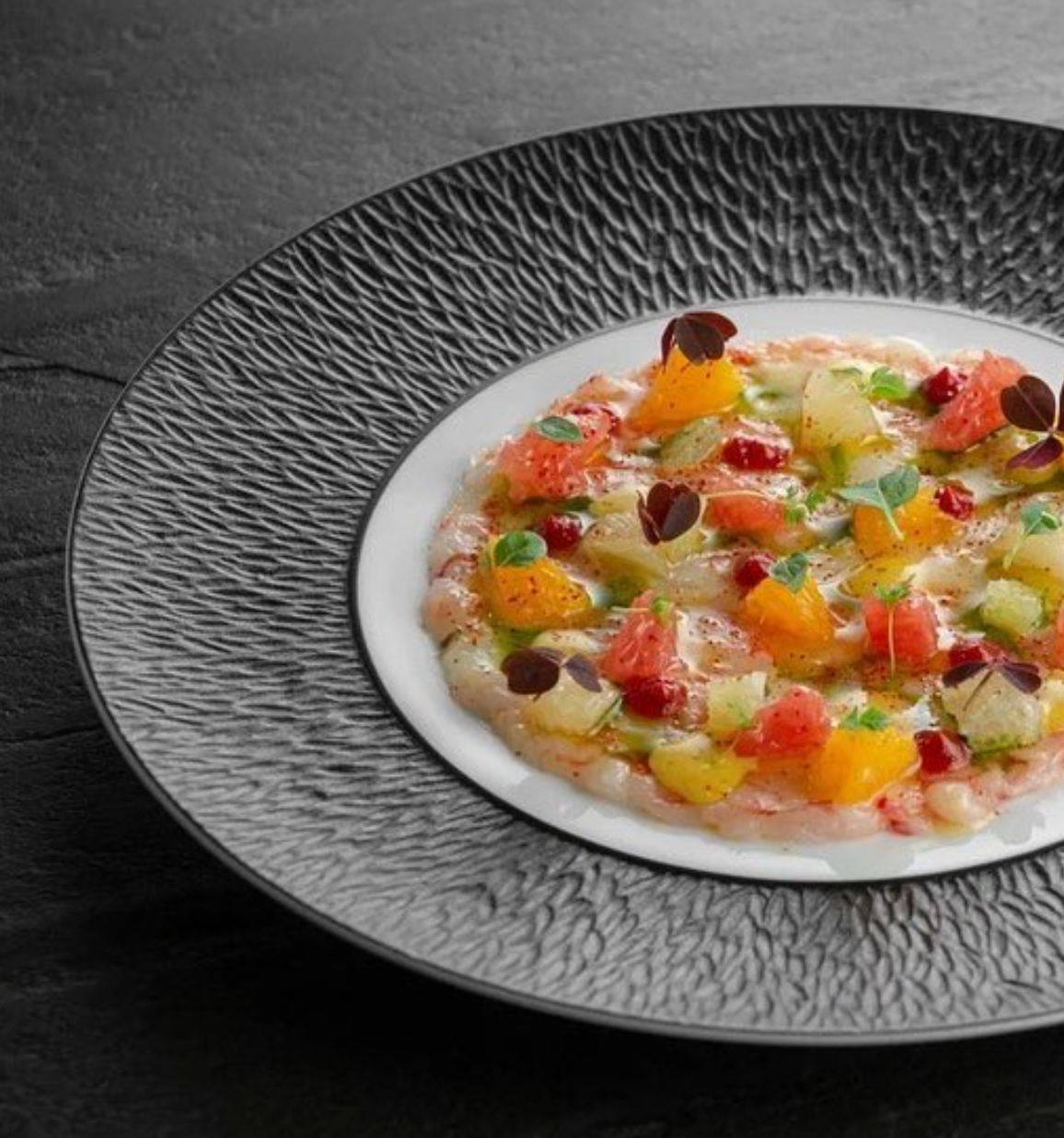 A gourmet dish is artfully presented on the Mineral Irise Dinnerware Dark Grey by Raynaud, with vibrant fruit and herb pieces atop a translucent base, garnished with microgreens for a striking, colorful display.