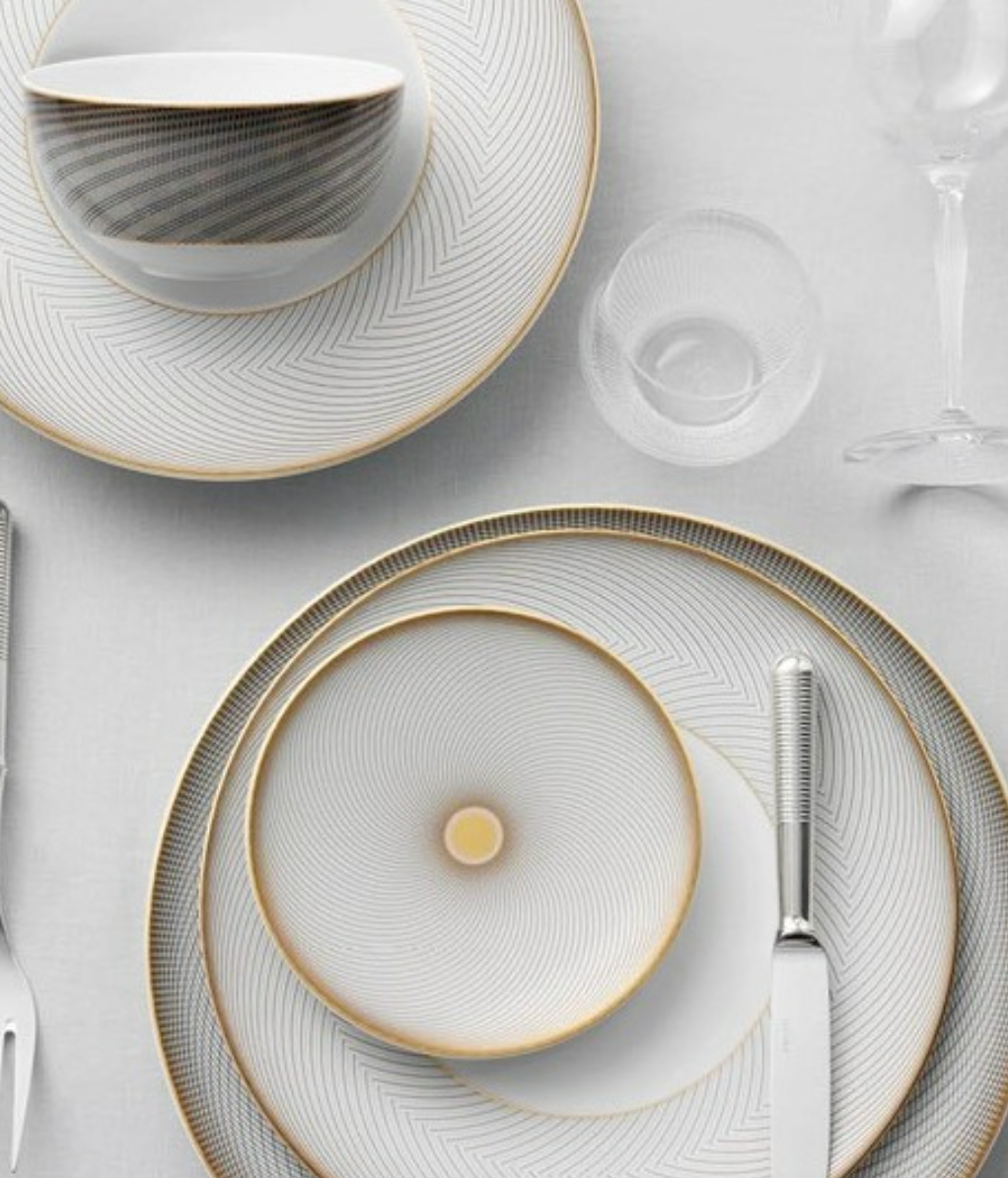 Elegant table setting with Oskar Dinnerware by Raynaud—white dishes with thin gold and gray geometric designs, plus a bowl, glass, fork, and knife on a white tablecloth—perfect for any occasion.