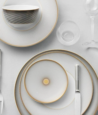 Elegant table setting with Oskar Dinnerware by Raynaud—white dishes with thin gold and gray geometric designs, plus a bowl, glass, fork, and knife on a white tablecloth—perfect for any occasion.