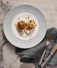 Two seared scallops on a creamy puree, garnished with chopped herbs and nuts, are served on a Raynaud Mineral Irise Dinnerware Pearl Grey plate, placed on marble beside a gray cloth napkin with a fork and knife.