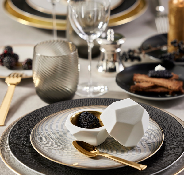 A geometric white Oskar Dinnerware dish by Raynaud with a lid holds blackberries atop a gold-accented plate. A gold spoon, fork, glassware, and crackers with cheese complete the elegant dining table setting.