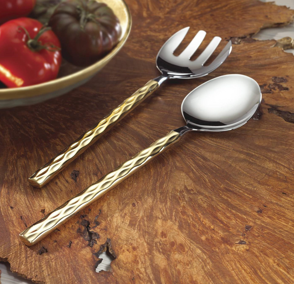 The Michael Wainwright Truro Salad Servers Gold, featuring 18/10 stainless steel and 24k gold textured handles, rest on a wooden surface beside a bowl of tomatoes.