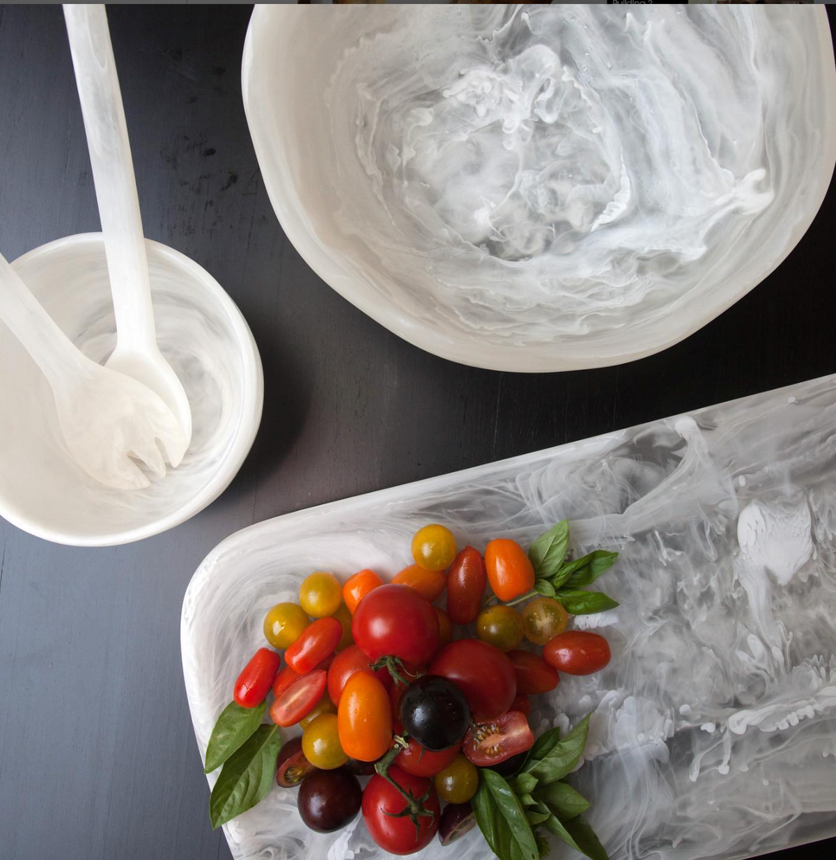 A marble platter with cherry tomatoes and basil sits next to the Nashi Home Swirl Resin Classic Wave Bowl and salad servers, all holding traces of creamy dressing atop a dark table.