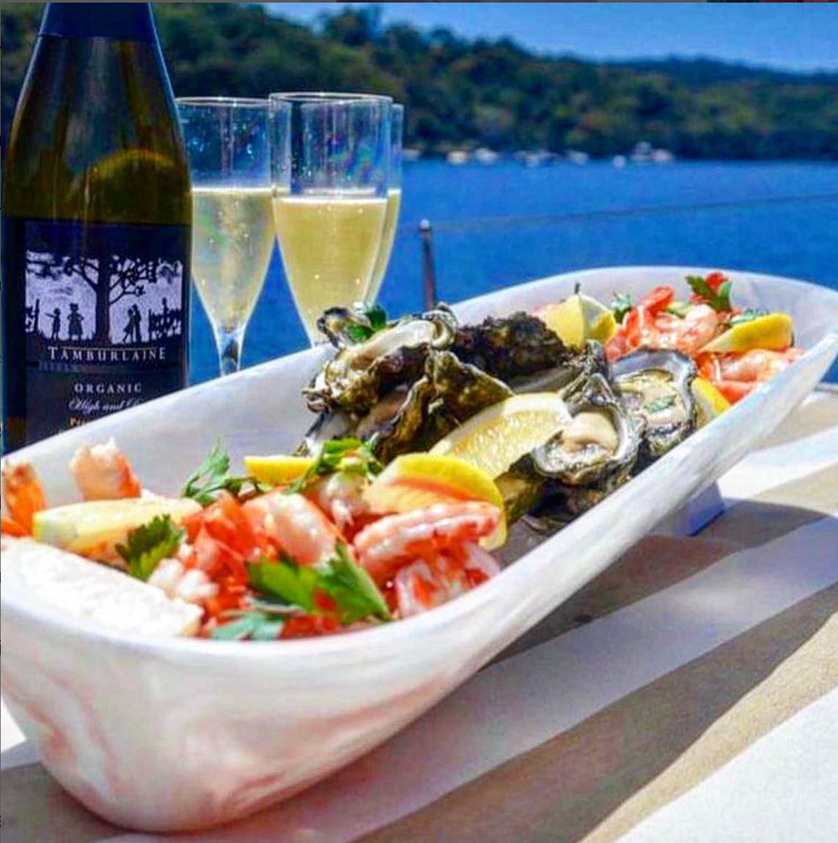 A Swirl Resin Classic Boat Bowl Jumbo by Nashi Home overflows with prawns and oysters plus lemon wedges, set beside two sparkling wine glasses and bottle on a table with scenic water views in the background.