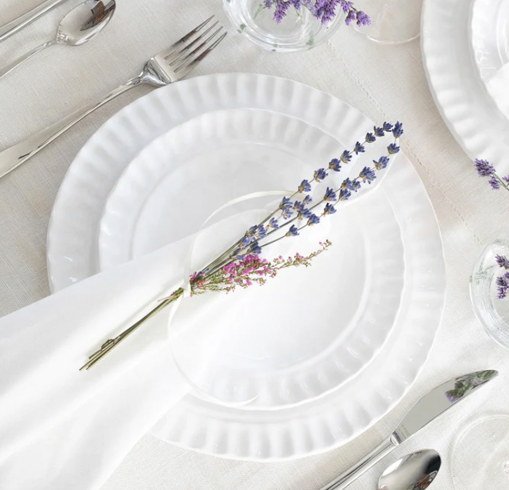 The Pietra Serena dinnerware set by Vietri is displayed on a white tablecloth with a cloth napkin topped by two sprigs of lavender. Silver cutlery and clear glasses encircle the plate, creating an elegant tablescape.