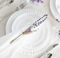 The Pietra Serena dinnerware set by Vietri is displayed on a white tablecloth with a cloth napkin topped by two sprigs of lavender. Silver cutlery and clear glasses encircle the plate, creating an elegant tablescape.