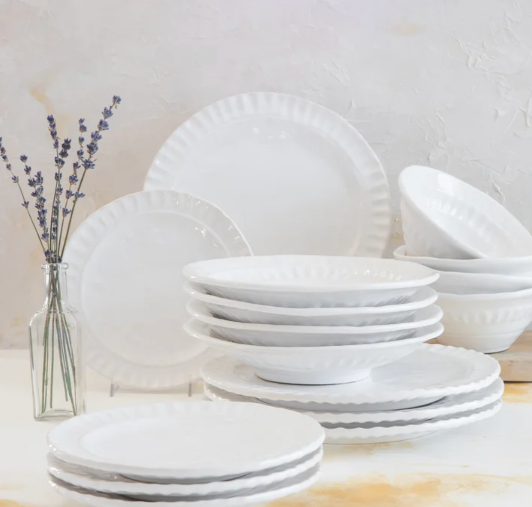 The Vietri Pietra Serena white dinnerware set, featuring plates and bowls with scalloped edges, is neatly stacked on a white surface beside a glass vase with lavender, set against a textured light background.