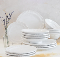 The Vietri Pietra Serena white dinnerware set, featuring plates and bowls with scalloped edges, is neatly stacked on a white surface beside a glass vase with lavender, set against a textured light background.