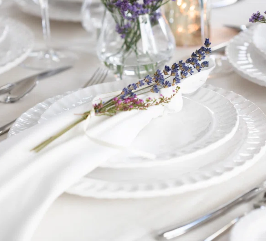 A Pietra Serena white dinner plate by Vietri, topped with a cloth napkin tied with lavender sprigs, is arranged among silver cutlery and glassware on a light tablecloth, with a vase of flowers in the background.