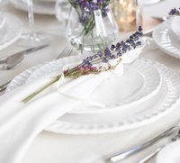 A Pietra Serena white dinner plate by Vietri, topped with a cloth napkin tied with lavender sprigs, is arranged among silver cutlery and glassware on a light tablecloth, with a vase of flowers in the background.
