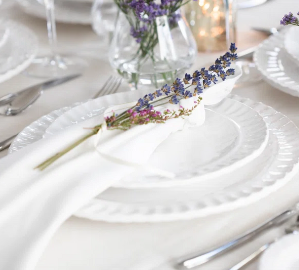 A Pietra Serena white dinner plate by Vietri, topped with a cloth napkin tied with lavender sprigs, is arranged among silver cutlery and glassware on a light tablecloth, with a vase of flowers in the background.