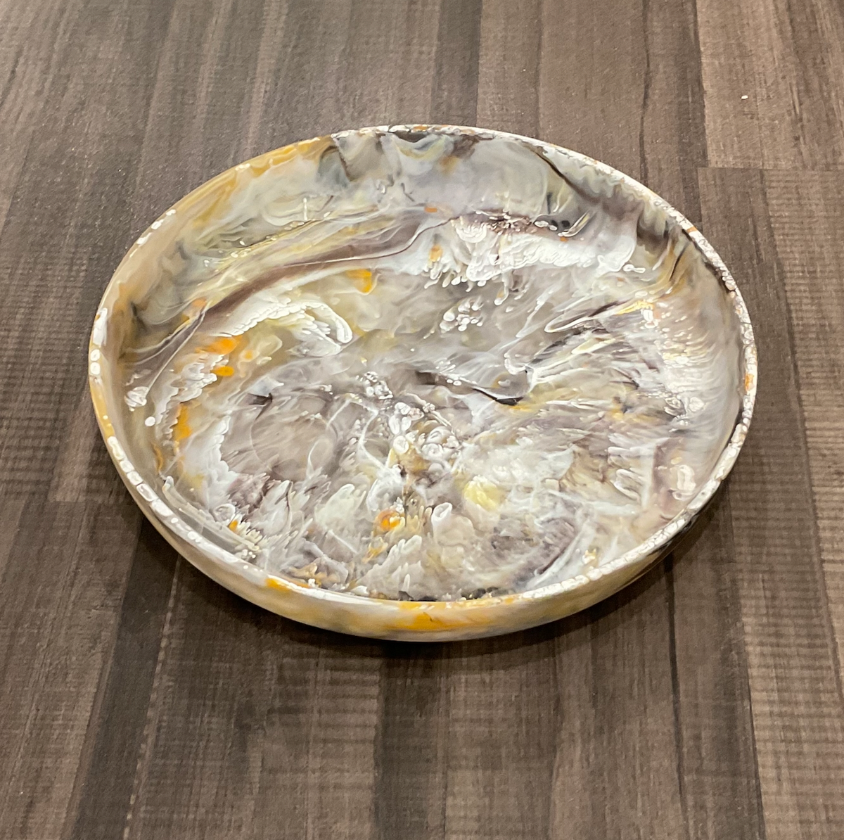 A round, shallow dish from Nashi Home—the Swirl Colored Resin Round Platter Mocha Medium—features a marbled pattern of gray, white, and accents of yellow and orange, and is placed on a dark wooden surface.