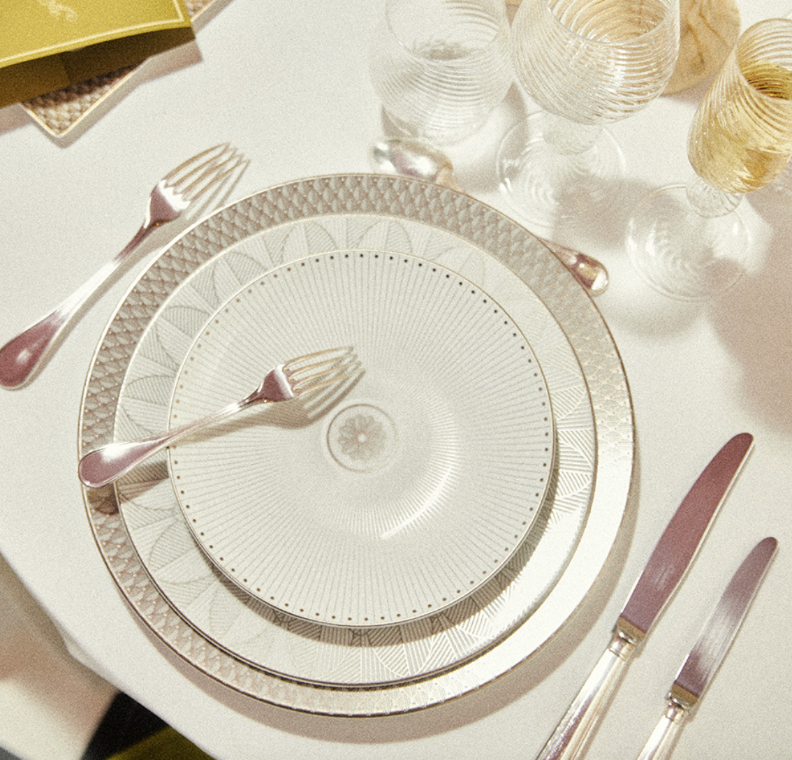 A formal table is set with Christofle Malmaison Imperiale Porcelain Dinnerware Platinum plates, a fork atop each dinner plate, two forks and a knife on the left, a knife and spoon on the right, and four textured glasses above.