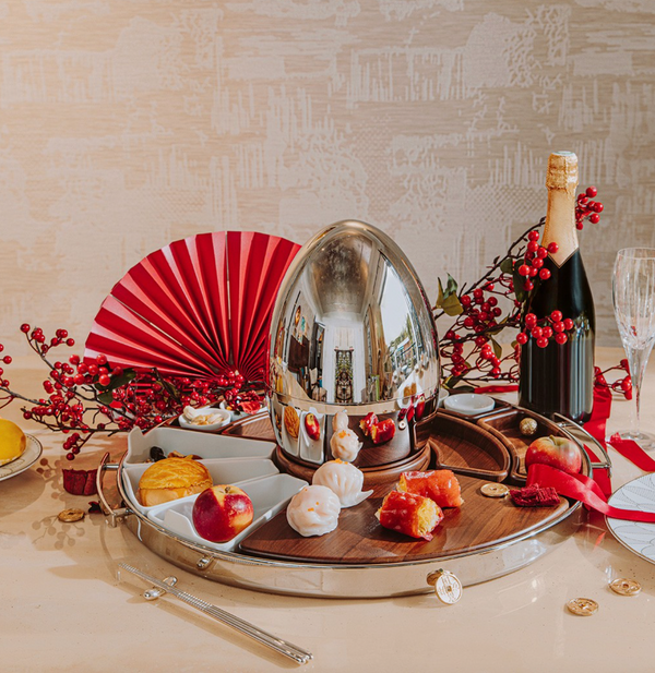 A Christofle MOOD Asia Tray – Stainless Steel is displayed with a shiny silver egg, assorted pastries, berries, a red paper fan, champagne, glasses, and gold coins on a table against a textured beige background.