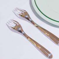 Two Christofle Infini Flatware silver-plated sporks from the 5 Piece Setting rest side by side on a white surface near a light green plate, with handles pointing downward.