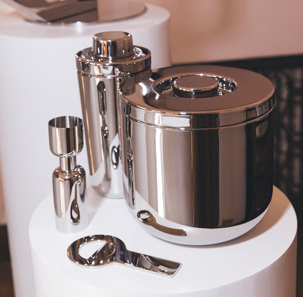 A set of four stainless steel bar tools—including the Christofle Oh De Insulated Ice Bucket, shaker, jigger, and bottle opener—from the Outdoor Series, elegantly arranged on a white circular stand.