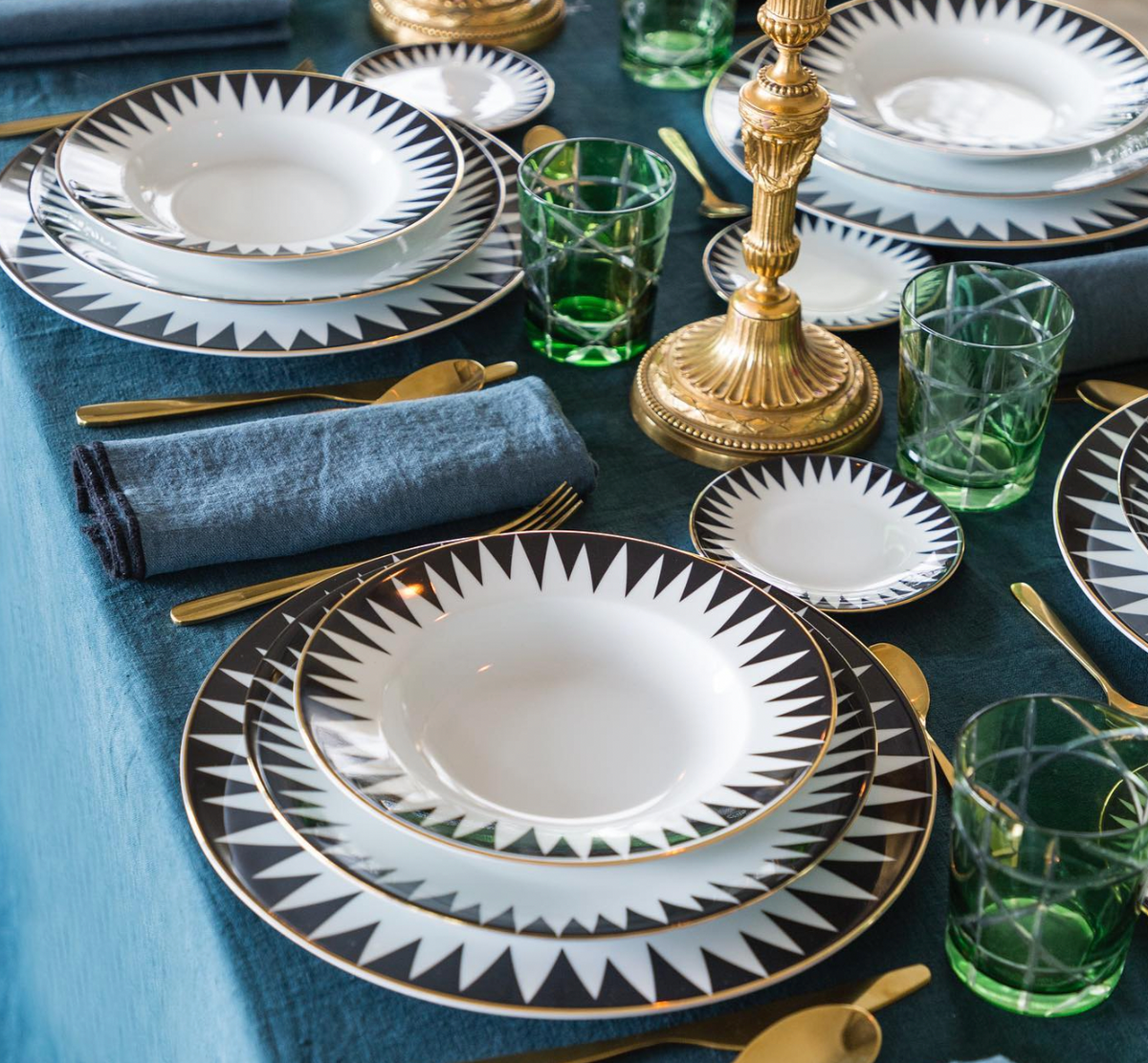 Elegant table set with Molecot’s Punk Dinnerware: white Limoges porcelain plates with black geometric patterns, gold flatware, green glass tumblers, a blue textured cloth, dark blue napkins, and a gold candlestick centerpiece.