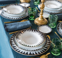 Elegant table set with Molecot’s Punk Dinnerware: white Limoges porcelain plates with black geometric patterns, gold flatware, green glass tumblers, a blue textured cloth, dark blue napkins, and a gold candlestick centerpiece.