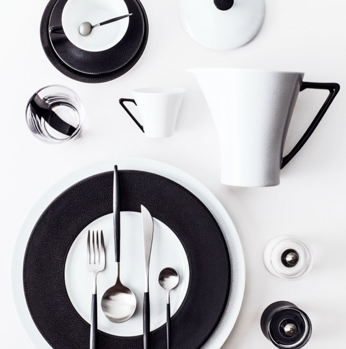 A modern place setting featuring the Philippe Deshoulieres Seychelles Black Charger 12.6", a white French porcelain dinner plate, cutlery, a cup, and a pitcher.