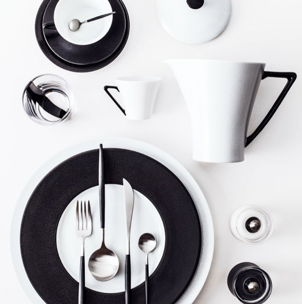 A modern, minimalist table setting showcases Philippe Deshoulieres’ Seychelles Dinnerware, featuring black and white plates with bold circular patterns, sleek cutlery, glassware, a jug, and a cup—all arranged on a clean white surface.