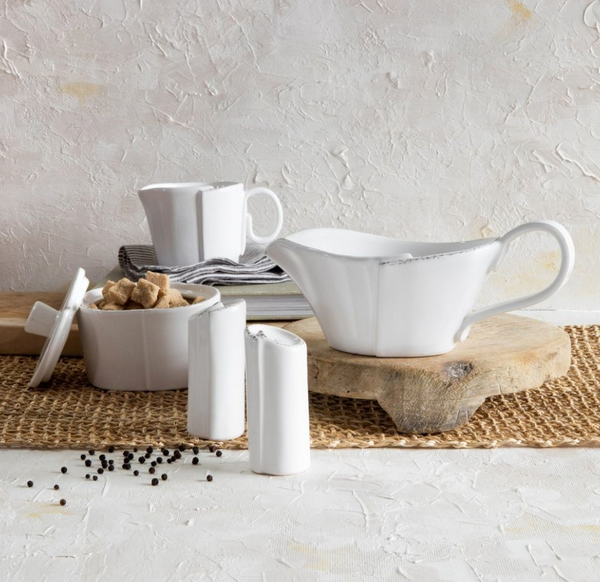 Vietri's Lastra Dinnerware White features a porcelain gravy boat with handle, sugar bowl with lid, salt and pepper shakers, and mug on a rustic surface accented by sugar cubes and peppercorns against a textured neutral background.