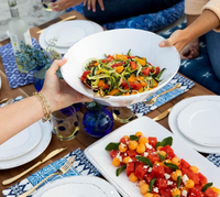 A hand holds a white bowl of zucchini noodle salad with tomatoes and peppers, next to a watermelon and cantaloupe salad on a stoneware platter—both elegantly served with Vietri Lastra Dinnerware White.