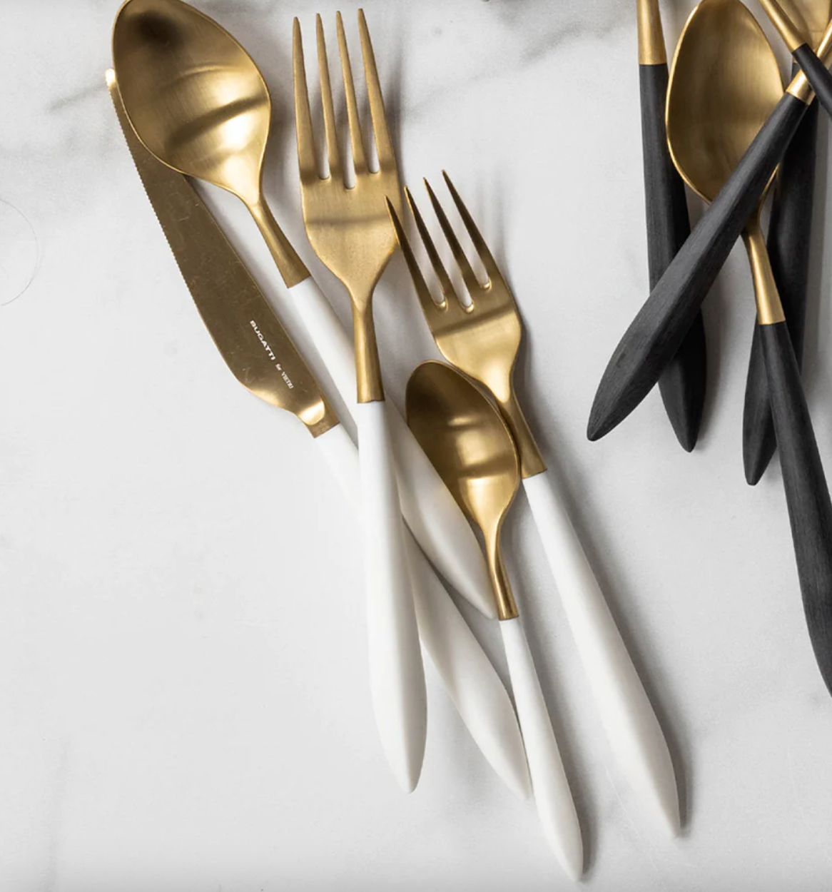 The Vietri Ares Flatware 5 Piece Setting in Oro & White features elegant gold and white stainless steel, part of a luxury Italian collection, with black-handled pieces in the background on marble.