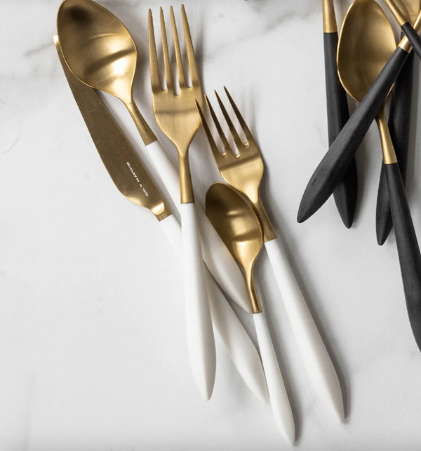 The Vietri Ares Flatware 5 Piece Setting in Oro & White features elegant gold and white stainless steel, part of a luxury Italian collection, with black-handled pieces in the background on marble.