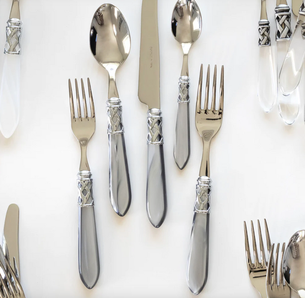 The Vietri Aladdin Brilliant Flatware 5 Piece Setting, featuring pearlized handles, forks, spoons, and a knife, is arranged on a white surface with additional 18/10 stainless steel utensils partly visible around the edges.