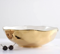 The PAMPA BAY Moonlight Bowl, a large glossy gold porcelain serveware piece with a white interior, is displayed on a white surface. Two dark cherries with stems are placed to the left. The background is plain white.