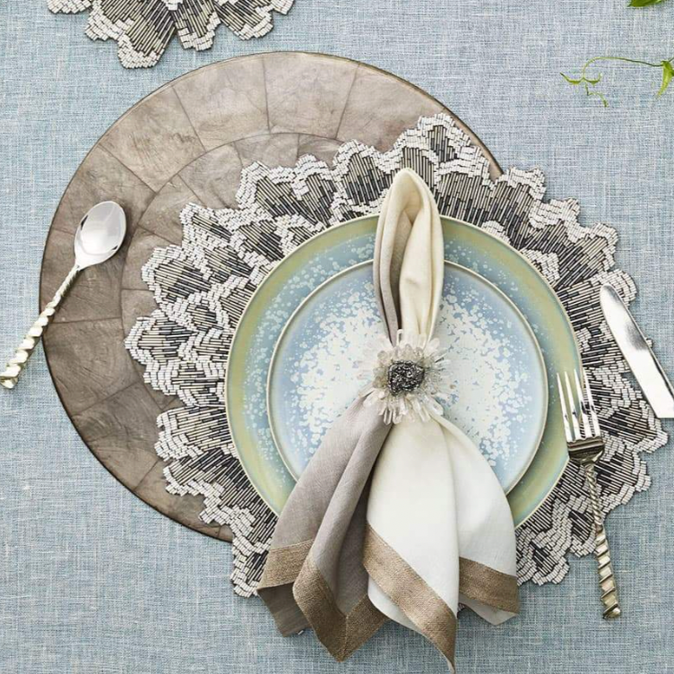 Elevate your table with layered plates, silverware, a folded napkin in a jeweled ring, atop a textured blue cloth and the Kim Seybert Capiz Shell Placemat Grey Set of 4 for extra sophistication.