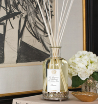 The Antica Farmacista/Harper Prosecco Diffuser, with its elegant label and long reeds, sits beside a white hydrangea in a vase on a table before framed art—a perfect home fragrance accent.