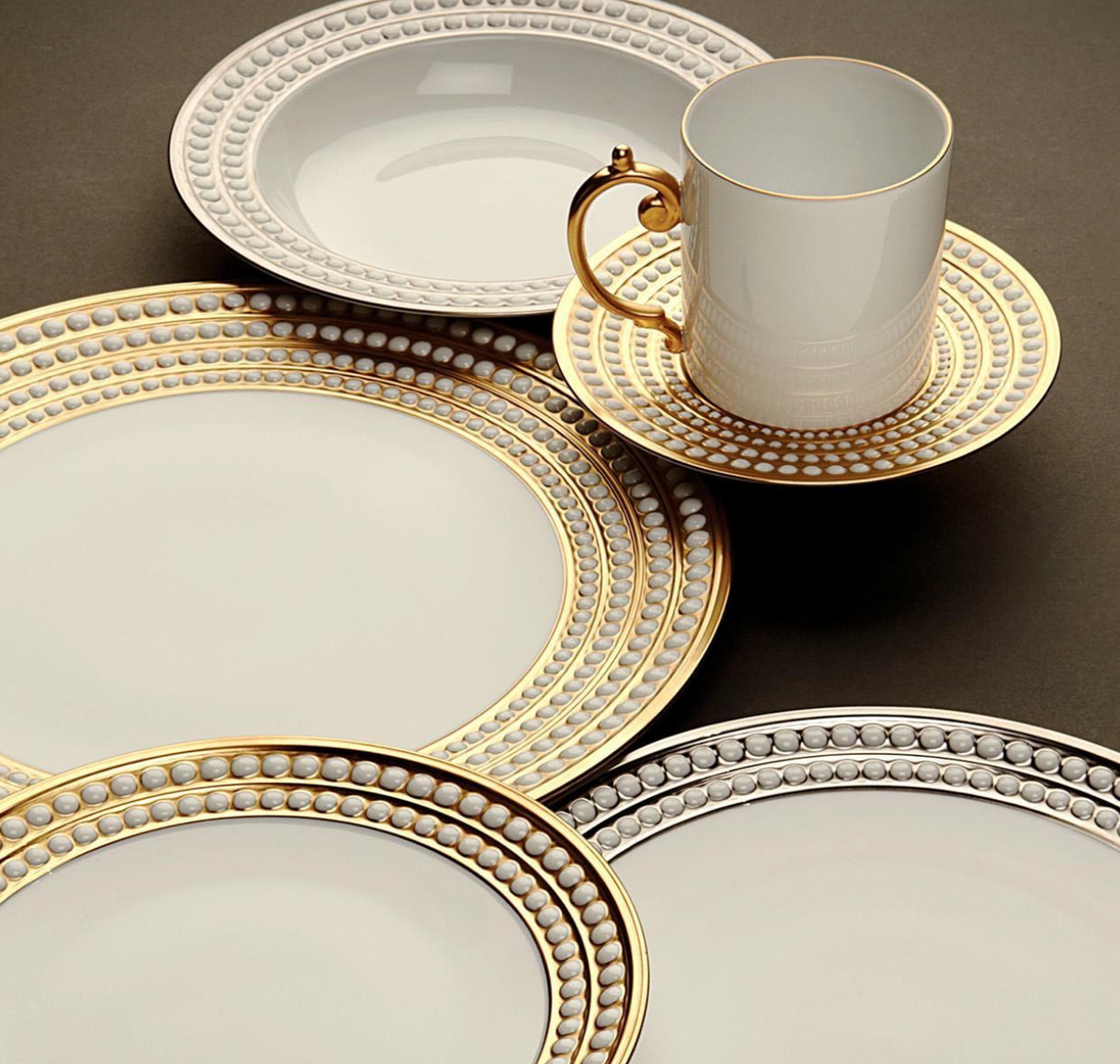 The L'Objet Perlée Platinum Dinnerware features elegant white porcelain plates with platinum rims, plus a cup and matching saucer. Ornate handles and signature accents add sophistication, beautifully displayed on a dark surface.