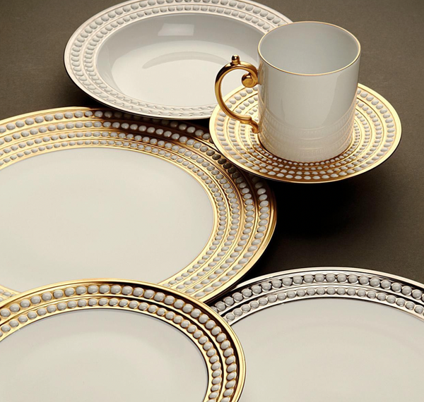 The L'Objet Perlée Platinum Dinnerware features elegant white porcelain plates with platinum rims, plus a cup and matching saucer. Ornate handles and signature accents add sophistication, beautifully displayed on a dark surface.