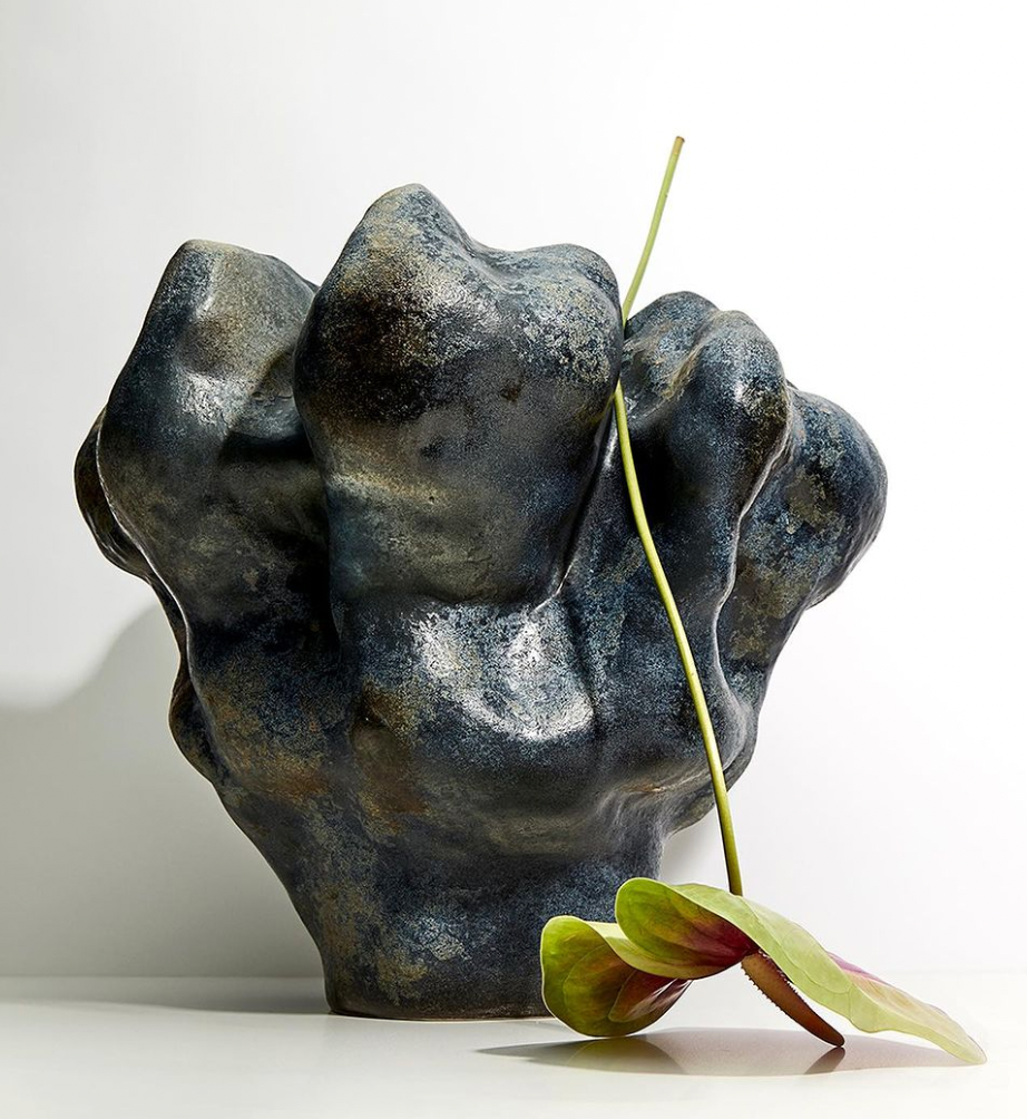The Timna Vase Short by L'Objet, featuring a dark, irregular form and rough texture, sits against a white background with a single long-stemmed flower with green and pink petals leaning across its surface.