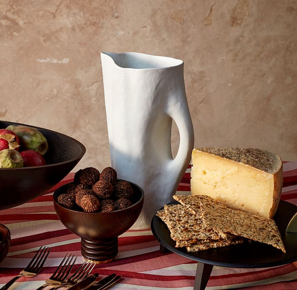 A Timna Porcelain Pitcher by L'Objet rests on a red and white striped tablecloth, surrounded by figs, walnuts, cheese, and seeded crackers on dark plates—an elegant piece of sculptural drinkware.