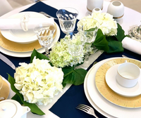 Elegant table setting featuring Haviland's Infini Prestige Gold Dinnerware, Limoges porcelain plates, crystal glasses, white napkins, and a centerpiece of white hydrangeas and green leaves on a navy blue and white tablecloth.