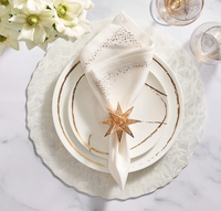 Elegant table setting featuring a white Sketch Chalk Dinnerware plate by Royal Crown Derby, a folded napkin with gold details in a gold star-shaped ring, clear glasses with gold rims, and white flowers in the corner.