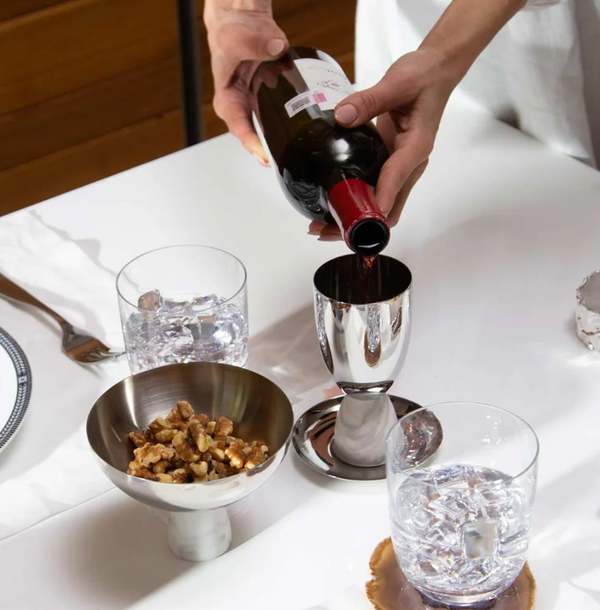 A person pours red wine from a bottle into the Anna New York Coluna Marble Kiddush Cup Silver on a white table set with glasses of ice water and a metal bowl of walnuts, accented by modern design touches.