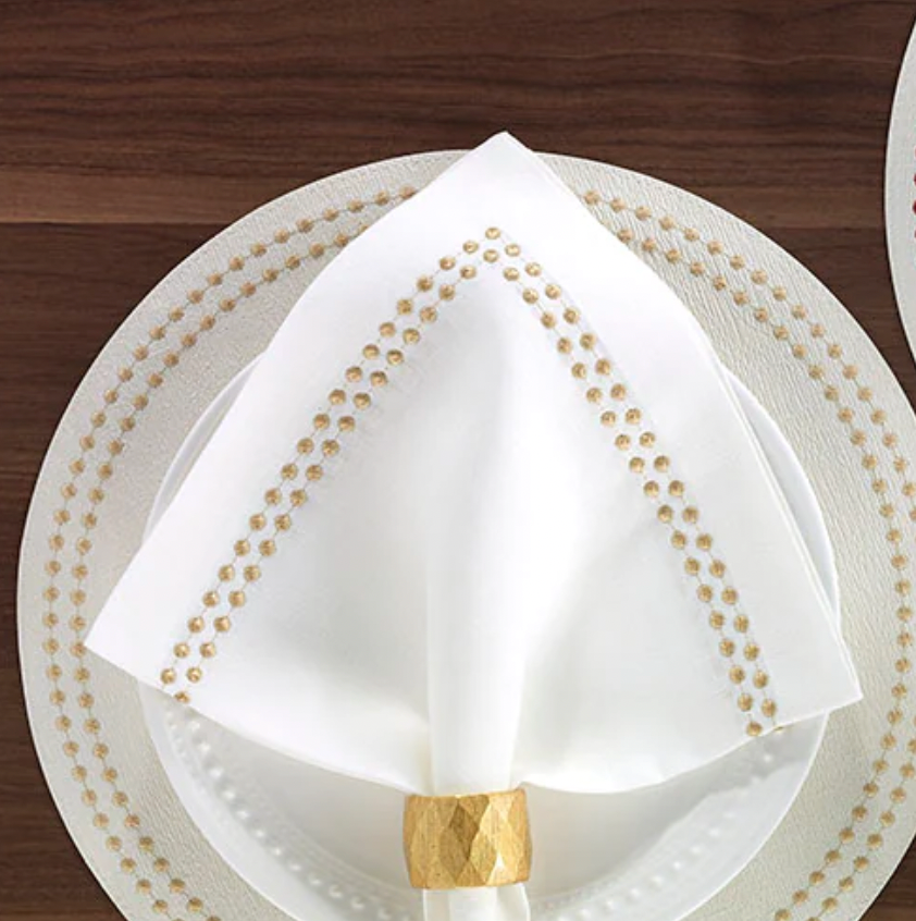 A Bodrum Pearls Napkin Gold, folded into a triangle and held by a gold ring, rests on a white plate atop a patterned charger on a dark wooden table.