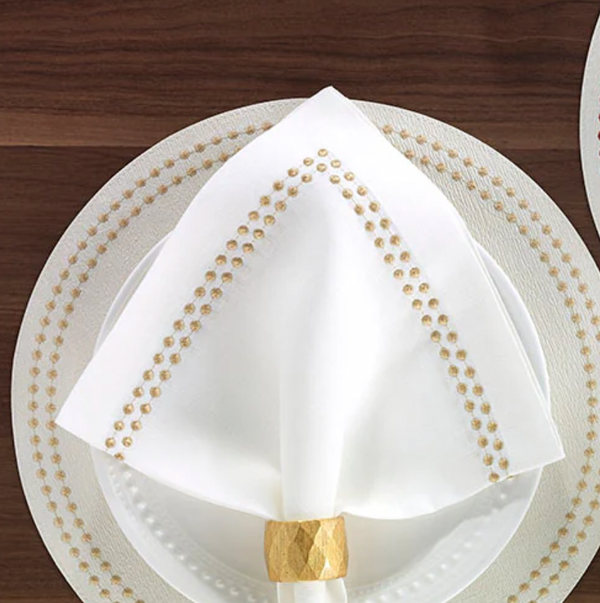 A Bodrum Pearls Napkin Gold, folded into a triangle and held by a gold ring, rests on a white plate atop a patterned charger on a dark wooden table.