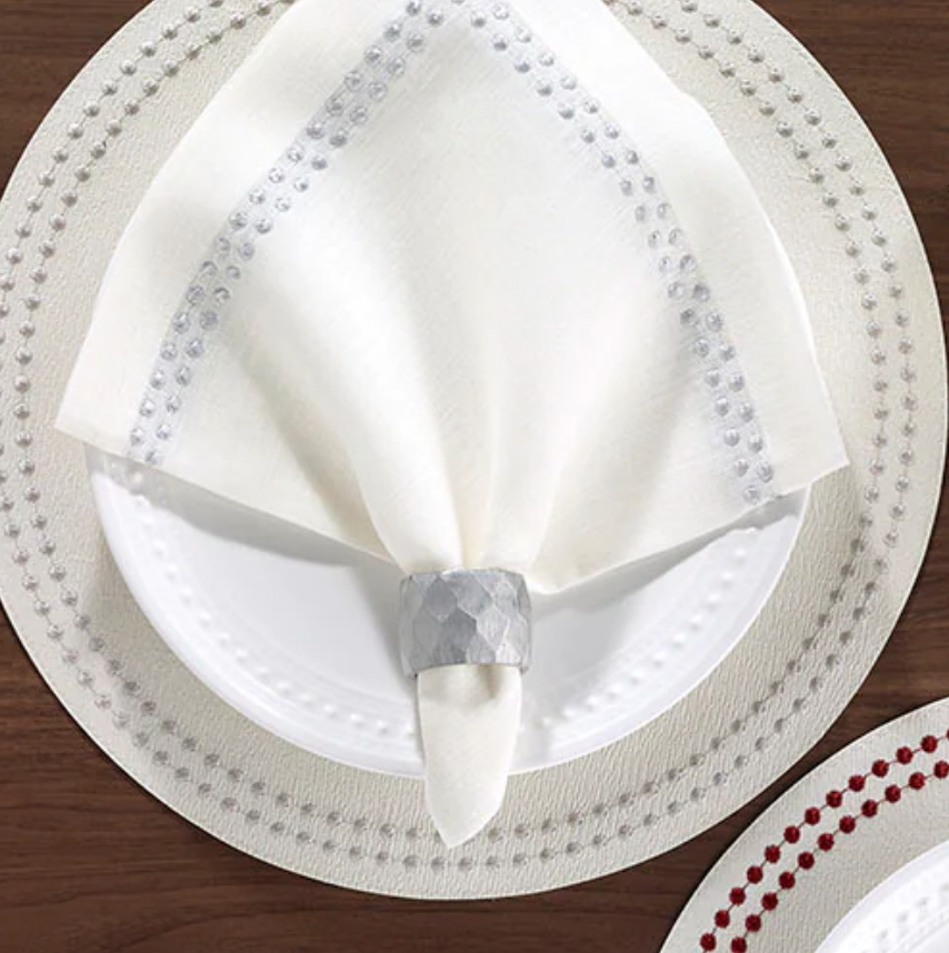 A white plate on a matching placemat features a Bodrum Pearls Napkin Silver Set of 4, finely crafted from linen and folded in a silver napkin ring, all elegantly arranged atop a wooden table.