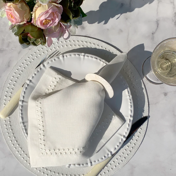 The Bodrum Pearls Napkin White Set of 4 is elegantly displayed on a textured placemat with a ring, accompanied by a knife, fork, glass of white wine, and floral arrangement atop a marble surface.
