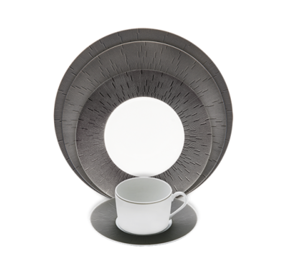 A white Limoges porcelain cup and saucer sit before a set of Haviland Anthracite Infini Dinnerware Grey plates, which feature textured radiating line patterns, all arranged on a white background.