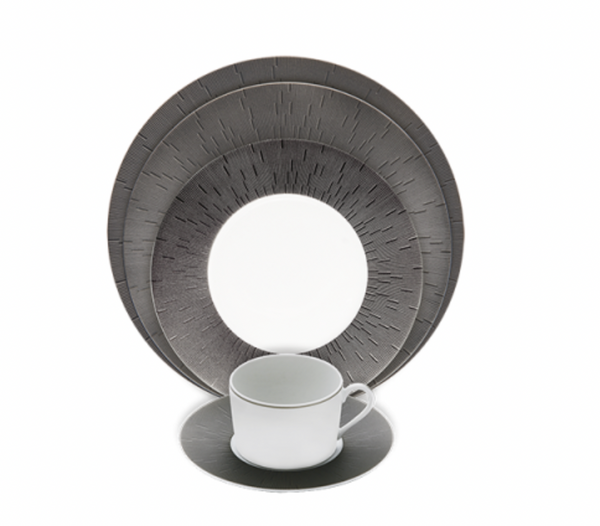 A white Limoges porcelain cup and saucer sit before a set of Haviland Anthracite Infini Dinnerware Grey plates, which feature textured radiating line patterns, all arranged on a white background.