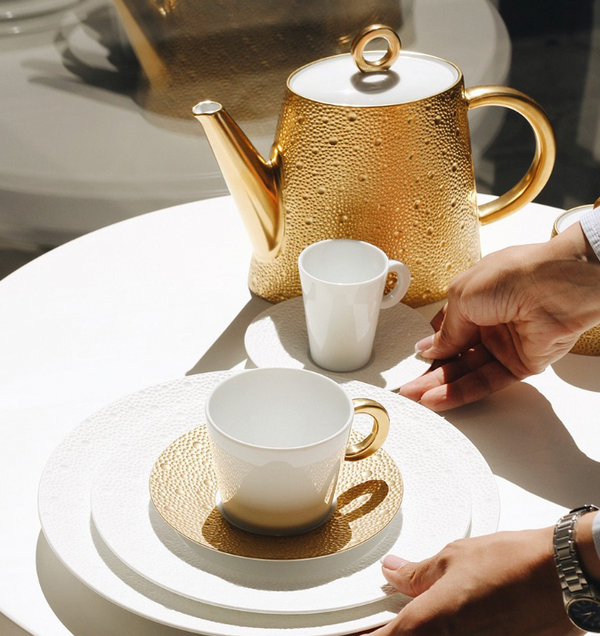 A person sets a textured white plate and gold-handled porcelain cup by the Bernardaud Ecume Gold Tea-Coffee Pot, with another cup, all in bright sunlight, showcasing the elegant Ecume dinnerware design.