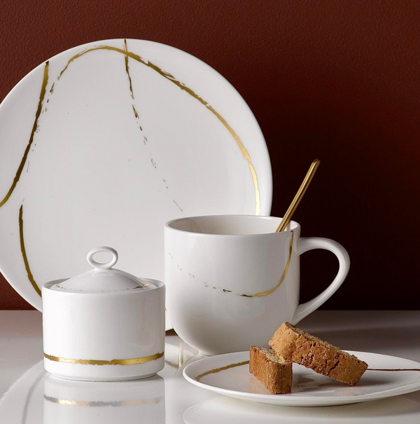 The Royal Crown Derby Sketch Chalk Dinnerware—a white plate, mug with gold spoon, and lidded sugar bowl, all English fine bone china with gold accents—is arranged on white. Two biscotti sit on a smaller plate before a brown background.