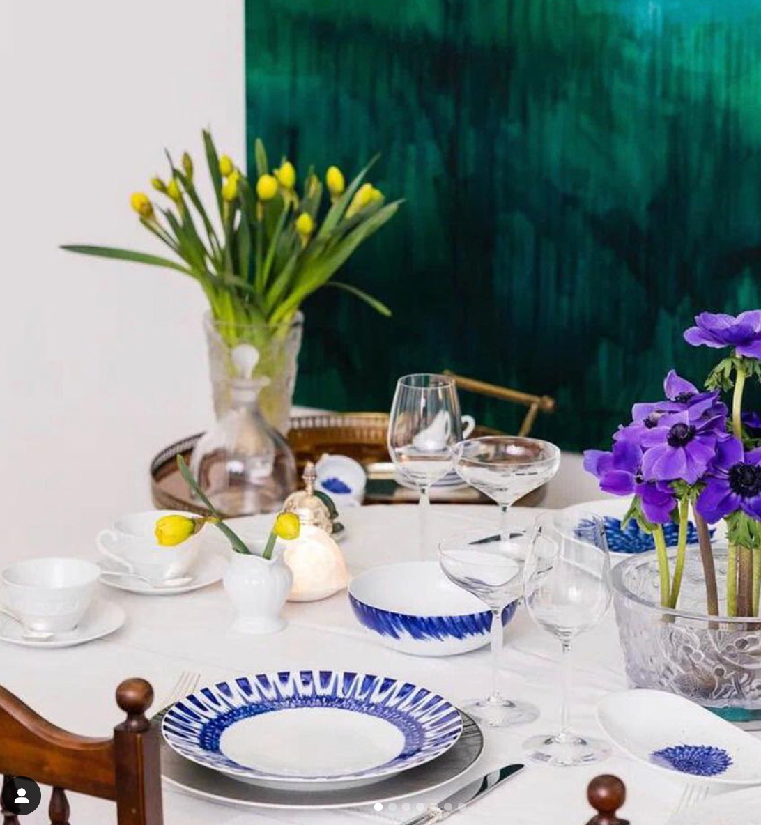A dining table set with blue and white In Bloom Dinnerware by Bernardaud and clear glassware. Yellow tulips in a vase and purple flowers add color, while a large green abstract painting hangs in the background.