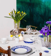 A dining table set with blue and white In Bloom Dinnerware by Bernardaud and clear glassware. Yellow tulips in a vase and purple flowers add color, while a large green abstract painting hangs in the background.